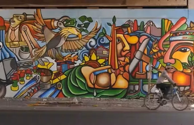Fresque street art colorée entremêlant des visages de personnages, des plantes, un aigle et des outils pour l'agriculture. Une personne passe en vélo devant la fresque.