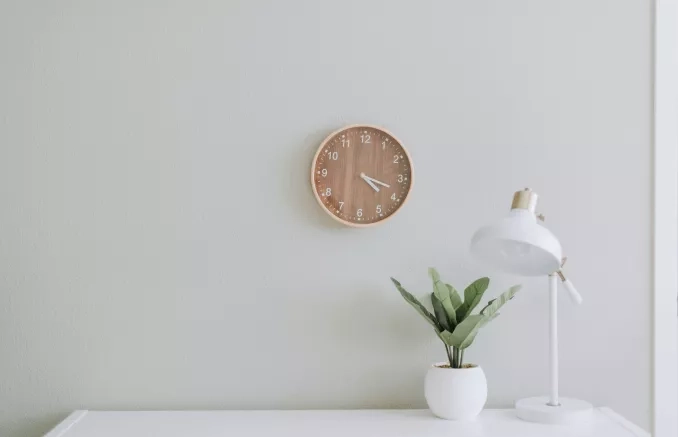 Photo d'un bureau très minimaliste avec une table blanche, une lampe de bureau blanche et dorée, un pot blanc avec une plante verte et une horloge murale en bois clair.